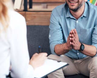 Ein Mann sitzt entspannt auf einem Sofa und spricht. Eine Person mit Notizen ist ihm gegenüber, die Atmosphäre wirkt freundlich und offen.
