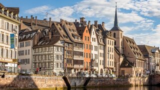 Das Bild zeigt malerische Fachwerkhäuser am Fluss in Straßburg, umrahmt von einem blauen Himmel mit einigen Wolken.