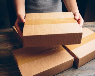 Eine Person hält ein braunes Paket in den Händen. Im Hintergrund liegen weitere Pakete auf einem Holztisch.
