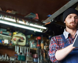 Ein Mann in Arbeitskleidung steht in einer Werkstatt voller Werkzeuge und nutzt ein Tablet. Die Werkstatt wirkt ordentlich und gut ausgestattet.