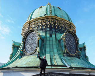 Eine Person steht oben auf einer detaillierten, grünen Kuppel mit großen, geschwungenen Fenstern. Der Himmel ist klar und blau.