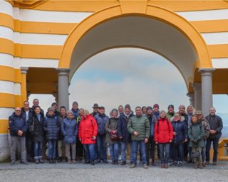 Eine Gruppe von Menschen steht unter einem großen, farbenfrohen Bogen. Sie tragen warme Kleidung und lächeln in die Kamera.