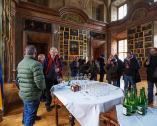 In einem prachtvollen Raum stehen Menschen zusammen, einige halten Gläser. Im Hintergrund sind beeindruckende Gemälde und eine festliche Tischdeko zu sehen.